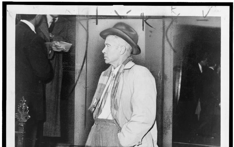 Black and white photograph of E.E. Cummings leaving a building---he is dressed smartly in some woven fabric trousers, a tartan scarf, and a hat.