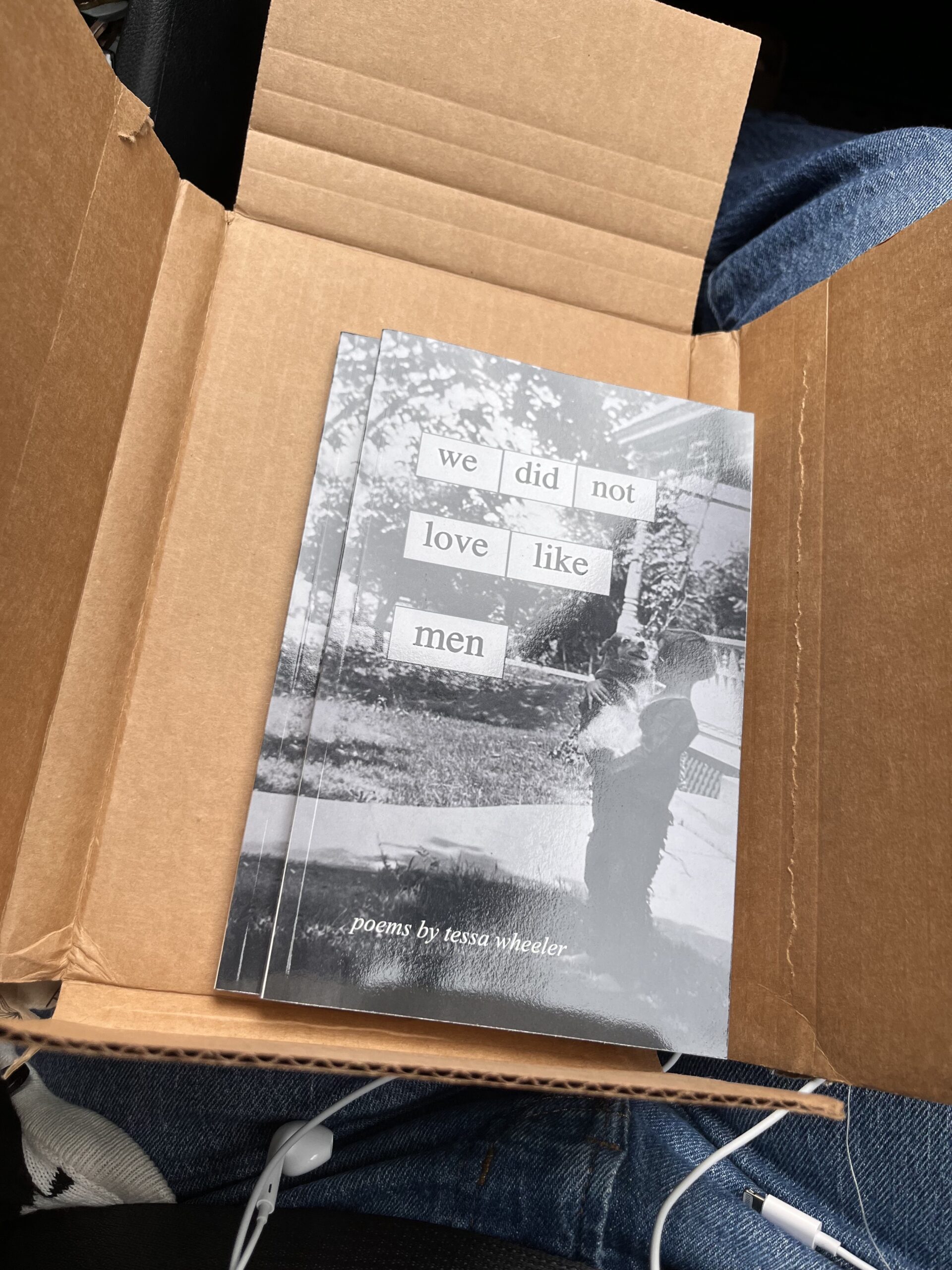 A cardboard envelope, opened, to reveal a stack of a few books inside. The cover says "we did not love like men", and has a black and white photo of a young girl holding a dog on it. At the bottom of the book, it says "poems by tessa wheeler".