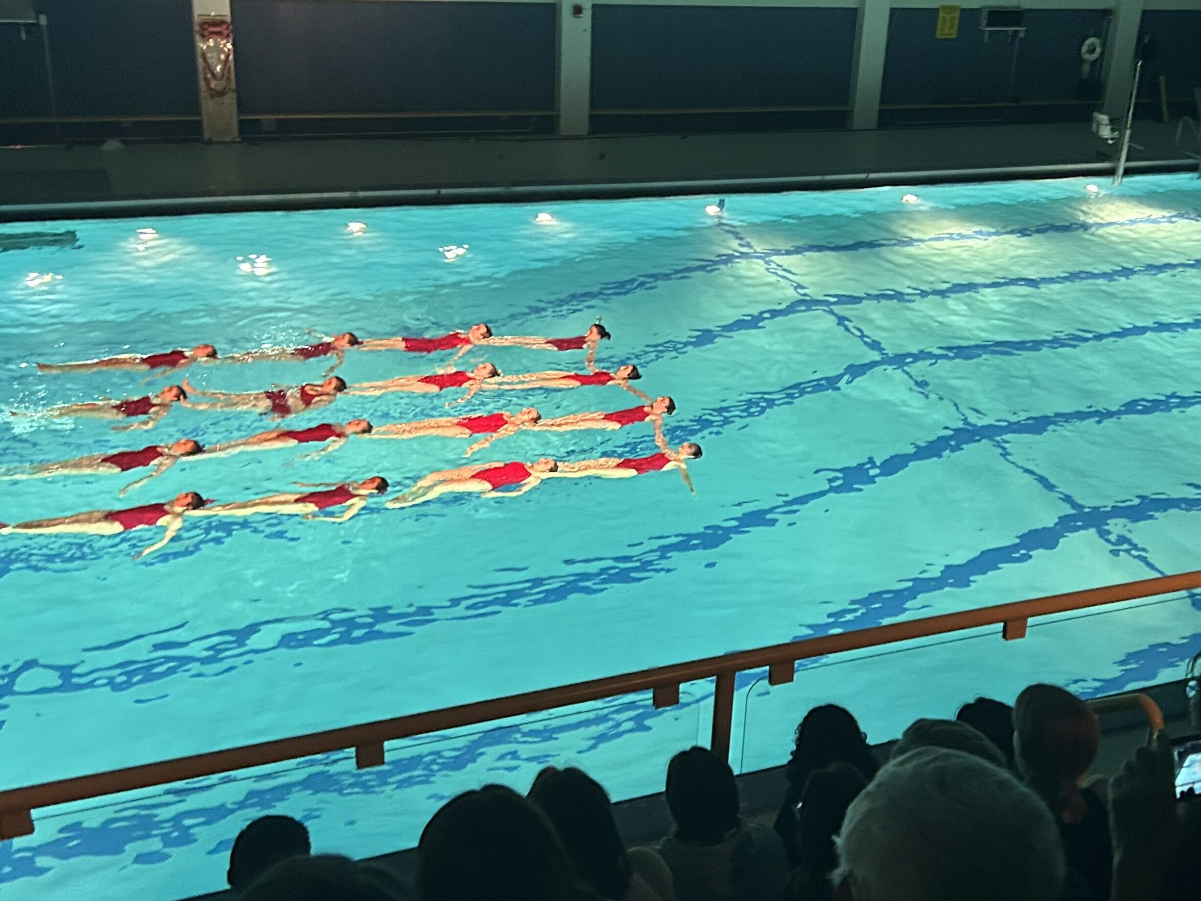 A color photograph of a bright blue pool seen from above. Audience members are seen in shadow taking phone photographs. In the water, sixteen synchronized swimmers in red swimsuits float on their backs in a 4x4 grid.