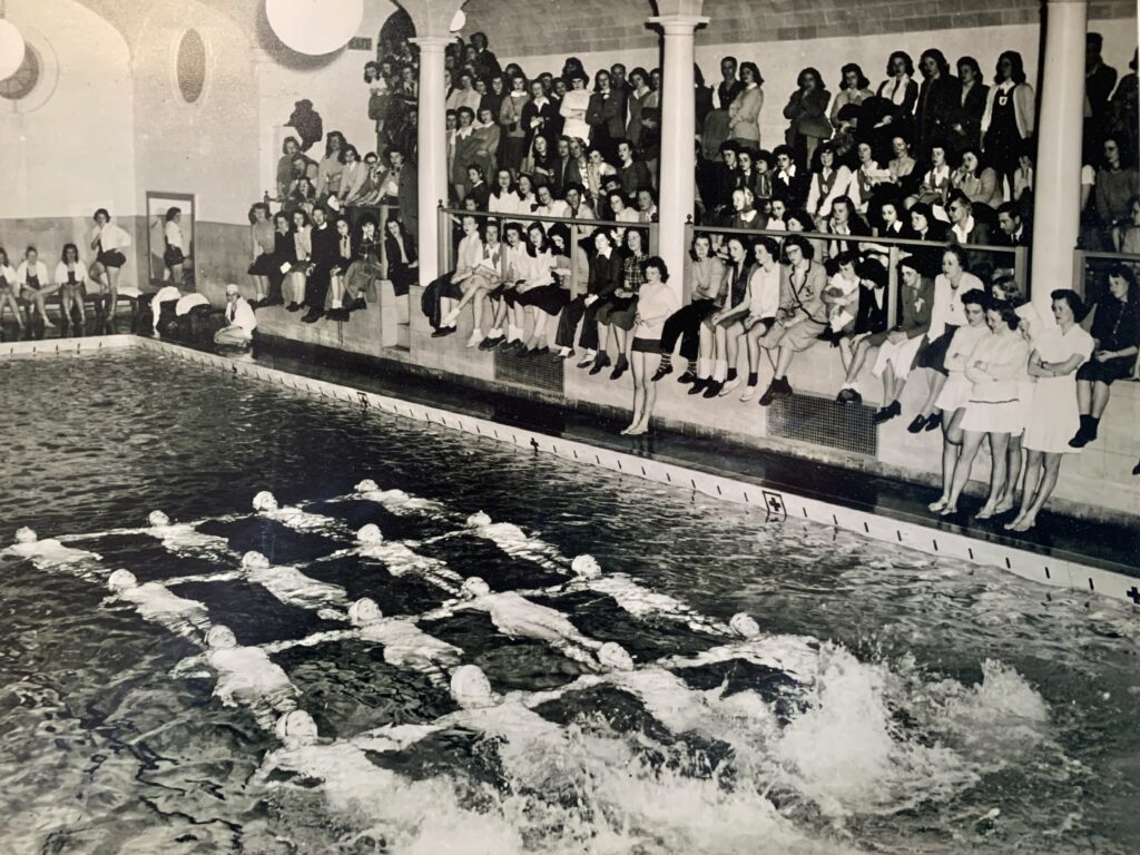 A black-and-white image of sixteen synchronized swimmers on their backs in a 4x4 grid. To their right, there is a tiered audience of Smith students in 1940s clothing.
