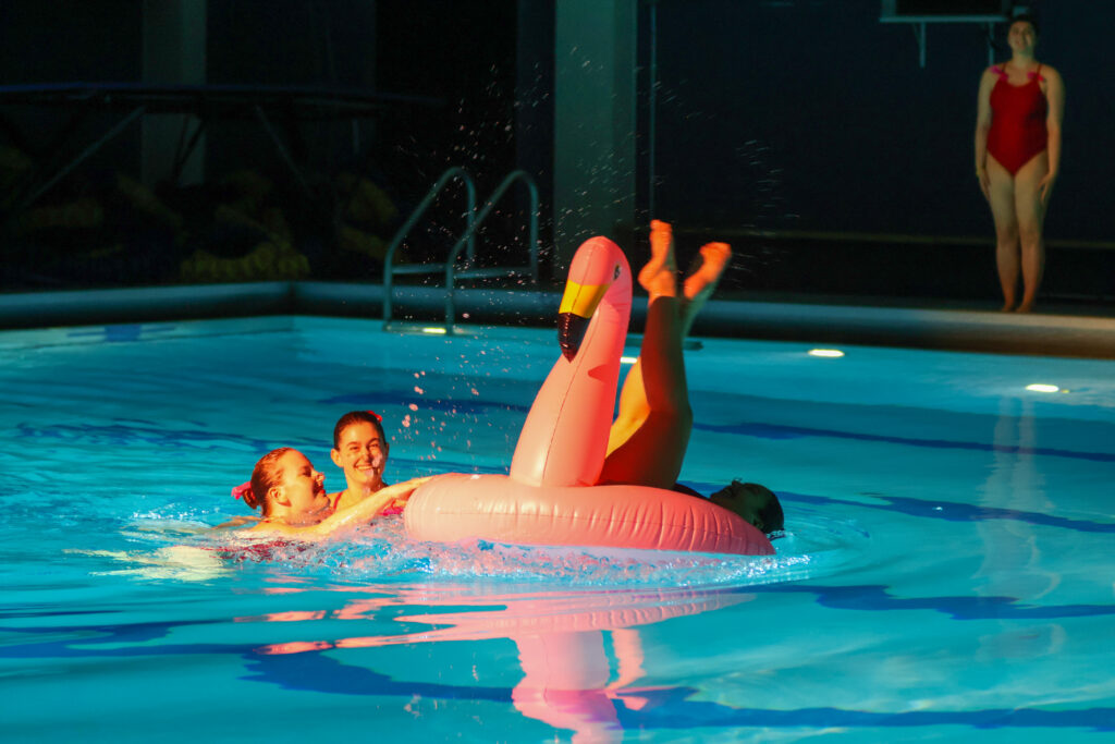 Two simmers push a pink blow-up flamingo-shaped float in a pool. Another swimmer falls out of the back of the float, feet in the air.