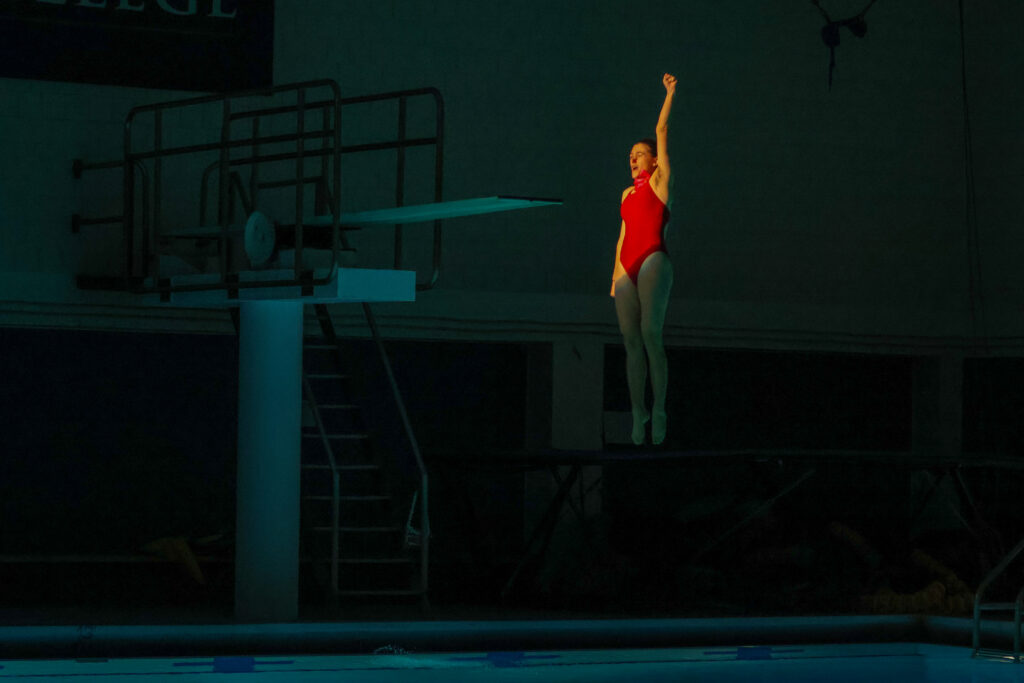 A swimmer in a red swimsuit free falling from the high diving board. She has one armed raised and is lighted by a spotlight.