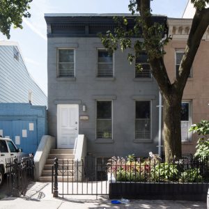Photo of blue, two-story row house with a white door, a small tree out front, and a small fence.